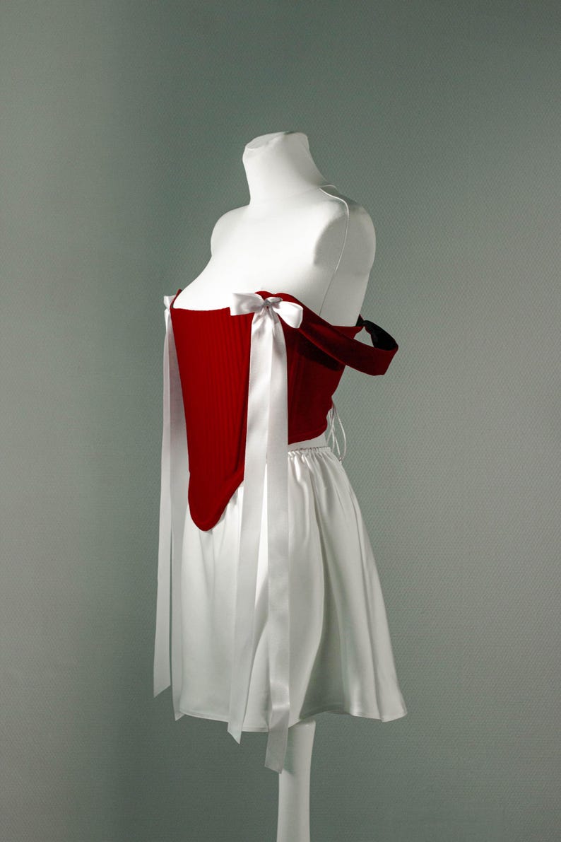 May include: A red corset-style top with white ribbon ties and a white skirt. The top has a structured design with a sweetheart neckline and is paired with a flowing skirt. The outfit is displayed on a mannequin.
