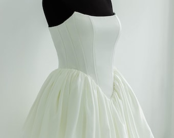 May include: A white strapless mini dress featuring a structured, corset-style bodice with vertical seams and a voluminous, gathered skirt that flares out from the waistline. The garment is displayed on a black mannequin torso.