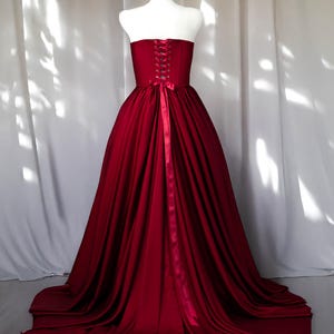 May include: A floor-length strapless gown in a deep burgundy red colour. The bodice features a corset-style back with matching ribbon lacing and a tied bow at the waist. The skirt is full and voluminous, cascading down to the floor in soft folds.