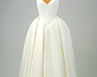 May include: A floor-length, strapless white gown featuring a structured sweetheart neckline and a fitted bodice. The skirt is voluminous with deep, vertical box pleats that extend from the waistline to the hem, creating a full, bell-shaped silhouette.
