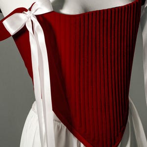 May include: A deep red, vertically-ribbed corset with white satin ribbon ties and bows. The corset is displayed on a white form, with a white skirt visible below. The overall style suggests historical or theatrical attire.