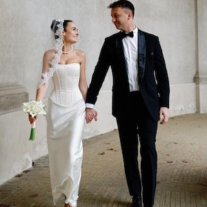 Puede incluir: Un vestido de novia blanco sin tirantes estilo corsé con acabado satinado y falda larga, junto a un velo con borde de encaje blanco y un ramo de calas blancas, al lado de un esmoquin negro con camisa blanca y pajarita negra.