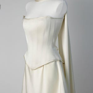 May include: An ivory-colored strapless corset top with vertical paneling and a pointed hemline, paired with a matching skirt. A long, flowing fabric piece is draped around the neck and extends down the back, creating a cohesive monochromatic ensemble.