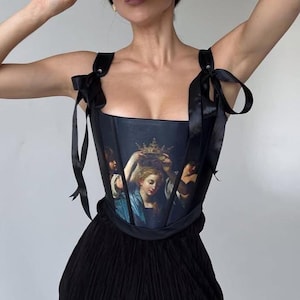 Puede incluir: Un corsé negro con una pintura de una mujer con una corona en la cabeza. El corsé tiene tirantes de satén negro que se atan en los hombros.