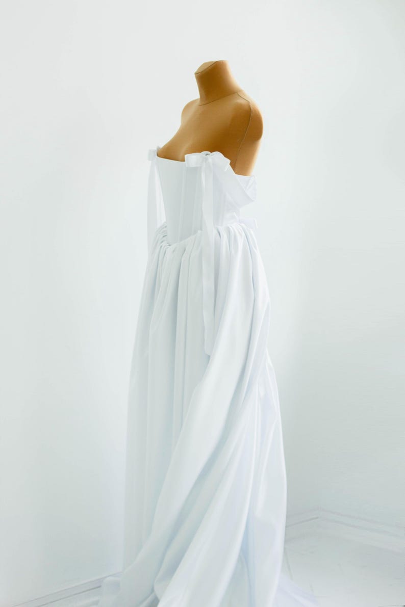 May include: A white, off-the-shoulder dress on a mannequin. The dress features a fitted bodice and a flowing skirt. The shoulders are adorned with delicate bows. The fabric appears to be lightweight and flowing, creating a soft, elegant silhouette.