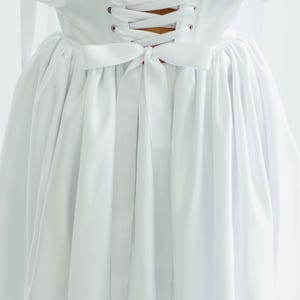 May include: A white dress with a corset back and a bow detail. The dress features a lace-up closure and a flowing skirt. The fabric appears to be a soft, lightweight material. The overall style is elegant and classic.