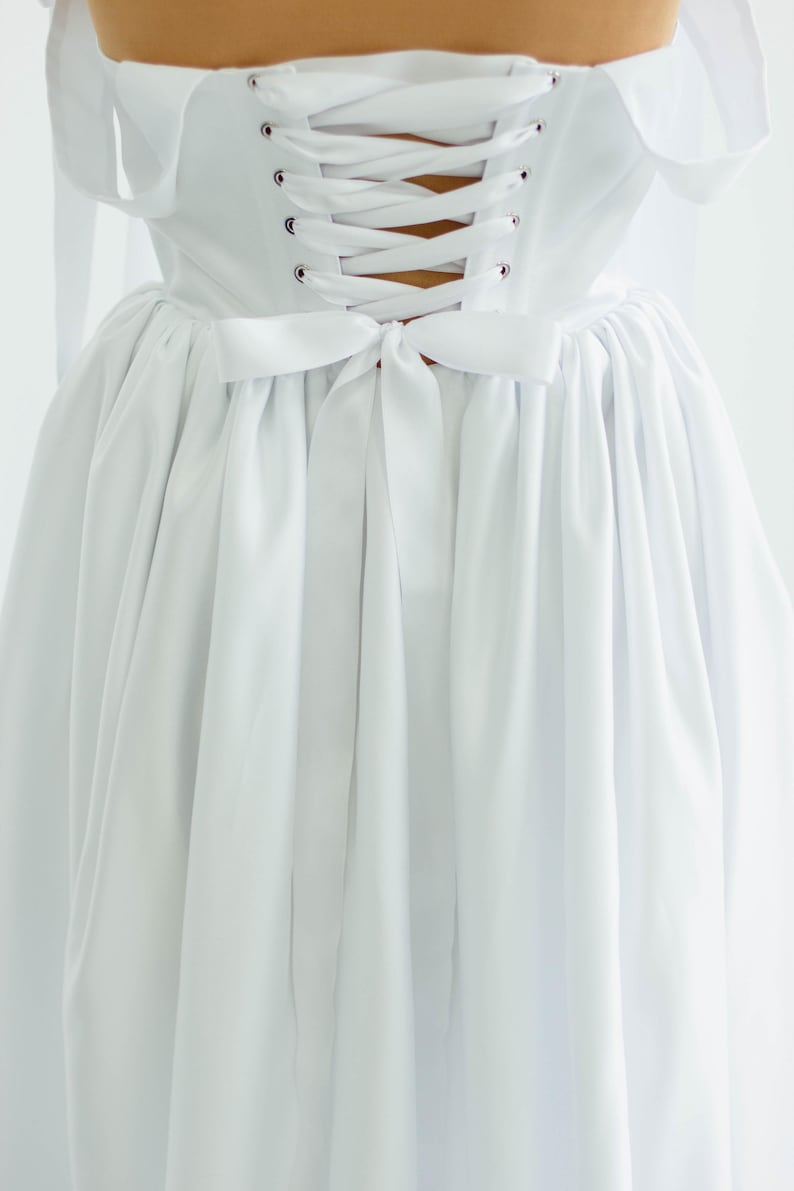 May include: A white dress with a corset back and a bow detail. The dress features a lace-up closure and a flowing skirt. The fabric appears to be a soft, lightweight material. The overall style is elegant and classic.