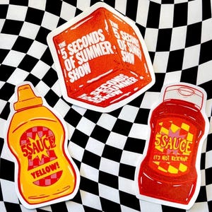 Puede incluir: Tres pegatinas troqueladas que incluyen un cuadrado naranja con el texto The 5 Seconds of Summer Show, una botella de mostaza amarilla con el texto 5SAUCE YELLOW y una botella de ketchup roja con el texto 5SAUCE IT'S NOT KETCHUP sobre un fondo de cuadros.