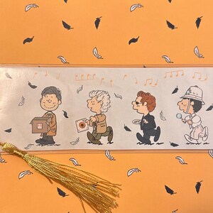May include: A rectangular laminated bookmark featuring four cartoon characters in a line, each holding a different object. A gold-colored tassel is attached to the left side. The background of the bookmark is white with small orange musical notes and feathers scattered around the figures.