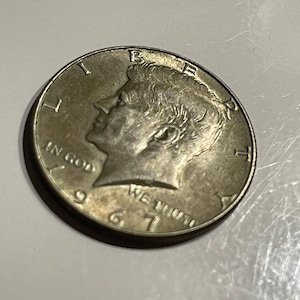 Könnte beinhalten: Eine silberne US-amerikanische Halbdolar-Münze mit dem Profil von John F. Kennedy auf der Vorderseite. Die Münze ist mit 1967 datiert und trägt die Inschrift "In God We Trust".