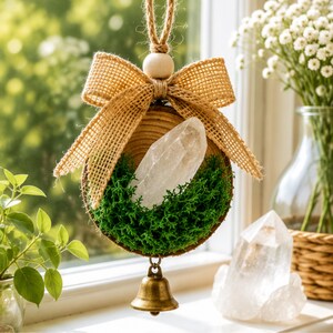 May include: A hanging ornament featuring a circular wood slice base topped with a large clear quartz crystal and vibrant green moss. A tan burlap bow and a wooden bead sit at the top, while a small brass bell hangs from the bottom of the wood.