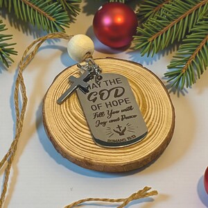 May include: A silver-colored dog tag pendant with a cross charm, engraved with the text "MAY THE GOD OF HOPE Fill You with Joy and Peace ROMANS 15:13". The pendant hangs on a wooden slice with a jute string and a wooden bead.