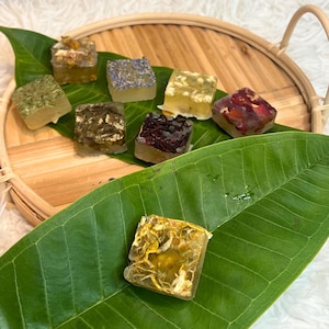 May include: A collection of square, clear soap bars infused with various dried flowers and herbs. The soaps are arranged on a wooden tray and a large green leaf. The soaps contain ingredients like lavender, rose petals, and chamomile.