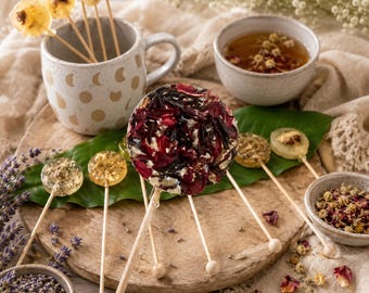 May include: A collection of round, clear hard candies on wooden sticks, featuring embedded dried flower petals and herbs. Some candies contain yellow blossoms, while one large central piece displays deep red and dark purple floral petals.