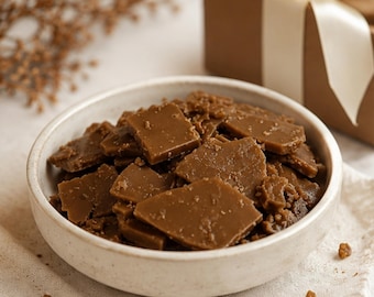 May include: A round, off-white ceramic bowl filled with irregular, flat, brown pieces of brittle candy. The pieces have a smooth, glossy surface with some granular texture. The bowl is positioned on a light-colored fabric surface.