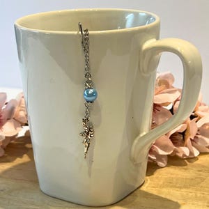 May include: A white ceramic mug with a silver tea infuser. The infuser has a chain with a light blue bead and a small fairy charm. Pink flowers are in the background.