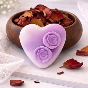 May include: A heart-shaped lavender soap bar featuring two raised rose embellishments on its surface. The soap is positioned in front of a dark wooden bowl filled with dried red rose petals, with additional loose petals scattered nearby.