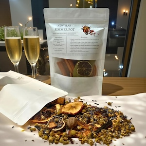 May include: A white pouch labeled New Year Simmer Pot containing dried orange slices, cinnamon sticks, and spices. The contents are spilled out, showing dried chamomile flowers, apple slices, nutmeg, and lavender. Two champagne flutes stand nearby.