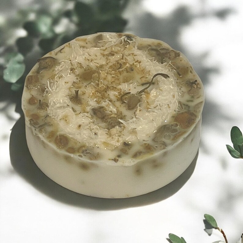 Op de afbeelding: Een ronde, witte zeep met een lichtbruine, getextureerde bovenkant. De zeep heeft gedroogde kruiden en bloemen erin verwerkt.