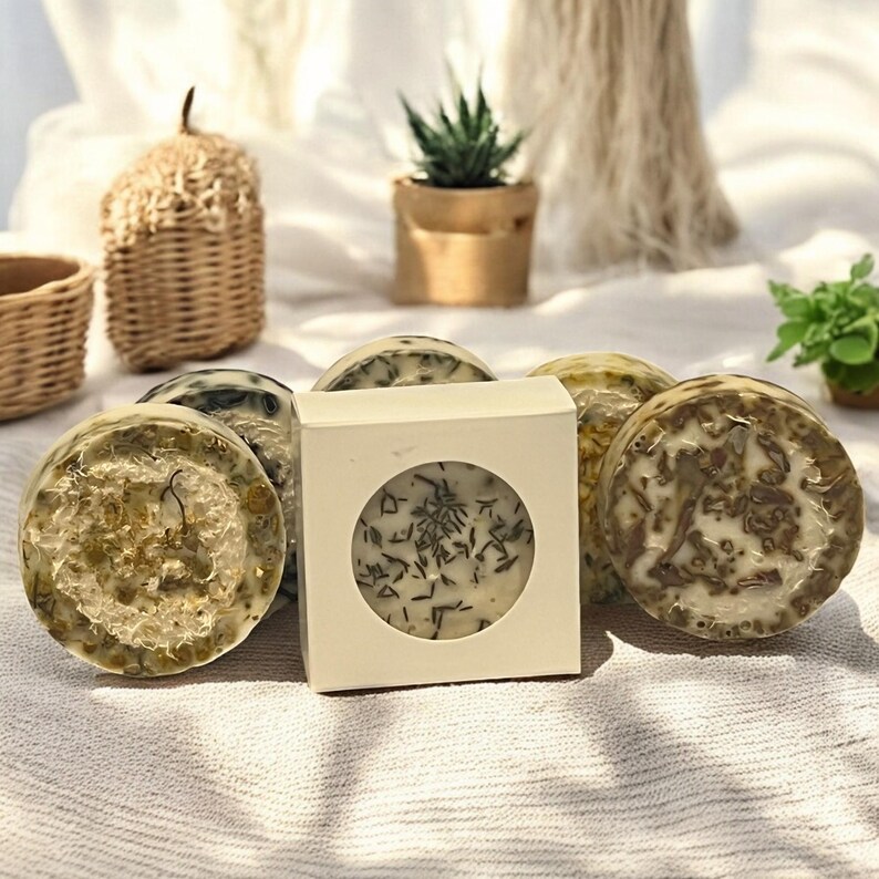 Op de afbeelding: Vijf ronde, witte zeepjes met groene en bruine botanicals erin. De zeepjes liggen op een wit stoffen oppervlak. E&eacute;n zeepje zit in een witte doos met een transparant venster waardoor je de zeep kunt zien.