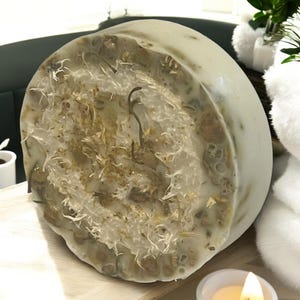 Op de afbeelding: Een ronde, witte zeepstaaf met een natuurlijke loofah die in het midden is ingebed. De loofah is lichtbruin van kleur en heeft een ruwe textuur. De zeepstaaf ligt op een houten oppervlak.