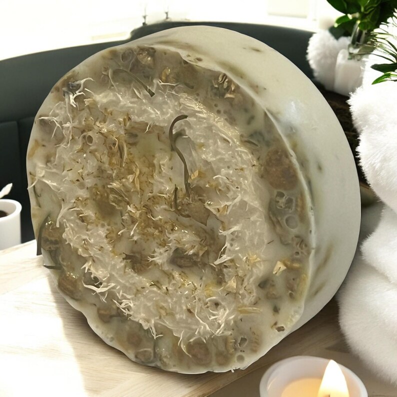 Op de afbeelding: Een ronde, witte zeepstaaf met een natuurlijke loofah die in het midden is ingebed. De loofah is lichtbruin van kleur en heeft een ruwe textuur. De zeepstaaf ligt op een houten oppervlak.