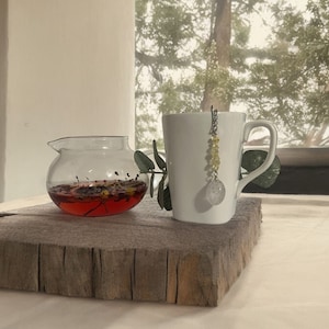 May include: A white ceramic mug with a handle and a glass tea infuser with a crystal charm and yellow beads hanging from it. The mug is sitting on a rustic wooden surface. The tea infuser is filled with red tea and flowers.