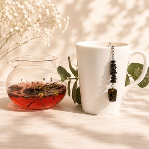 May include: A clear glass teapot filled with red herbal tea and dried flowers sits beside a white ceramic mug. A metal tea infuser hangs from the rim of the mug, featuring a chain of small black stone beads and a larger rectangular tiger eye stone.