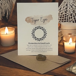 May include: A white rectangular card featuring the text Tiger Eye, a black mandala graphic, and the text Protection & Good Luck with a descriptive paragraph. A small, smooth, brown stone rests on a brown envelope positioned in front of the card.