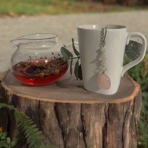 May include: A clear glass teapot filled with loose leaf tea sits on a tree stump next to a white ceramic mug with a rose quartz tea infuser.