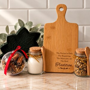 May include: A bamboo cutting board with the text The Recipe is Only The Beginning. The Memories are The Real Tradition. Two glass jars with cork lids, one filled with white powder and one with spices, and a clear ornament filled with spices.