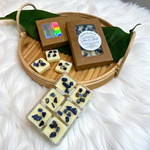 May include: Wax melts in a clear plastic container and cardboard boxes, arranged on a woven tray with a green leaf. The wax melts are square and cream-colored, topped with blue stones. The boxes have a clear window showing the wax melts.