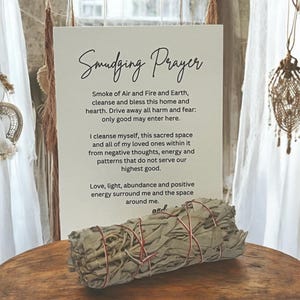 May include: A white rectangular sign with black text titled Smudging Prayer, featuring a cleansing invocation. In front of the sign sits a bundle of dried sage leaves wrapped in thin red string, resting on a circular wooden surface.