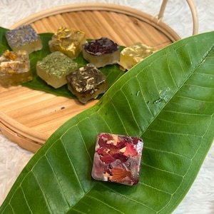 May include: A collection of square soaps with various botanical inclusions, such as flower petals and herbs. The soaps are displayed on a wooden tray and a large green leaf. The soap with red petals is in the foreground.