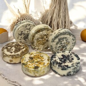 Op de afbeelding: Zes ronde, witte zeepstaven met verschillende natuurlijke ingredi&euml;nten en kleuren. De zepen zijn gerangschikt op een wit stofoppervlak.