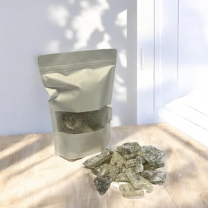 Pu&ograve; includere: Un sacchetto di carta bianco con una finestra trasparente che mostra un mucchio di caramelle dure verdi e bianche. Il sacchetto &egrave; appoggiato su una superficie di legno marrone chiaro. Un mucchio di caramelle dure verdi e bianche &egrave; sparso sulla superficie.