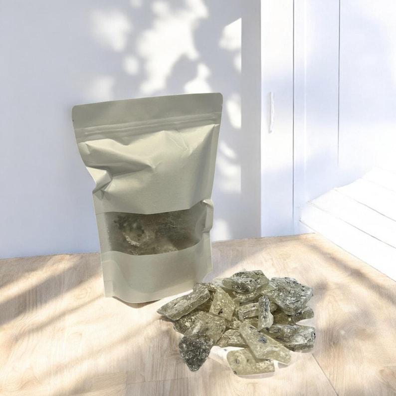 Pu&ograve; includere: Un sacchetto di carta bianco con una finestra trasparente che mostra un mucchio di caramelle dure verdi e bianche. Il sacchetto &egrave; appoggiato su una superficie di legno marrone chiaro. Un mucchio di caramelle dure verdi e bianche &egrave; sparso sulla superficie.