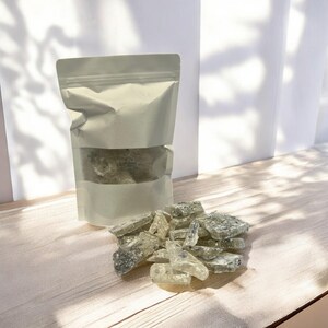 Pu&ograve; includere: Un sacchetto bianco richiudibile pieno di pezzi di caramelle trasparenti, verdi e bianche. Il sacchetto &egrave; appoggiato su una superficie di legno chiaro con un mucchio di pezzi di caramelle sparsi davanti.