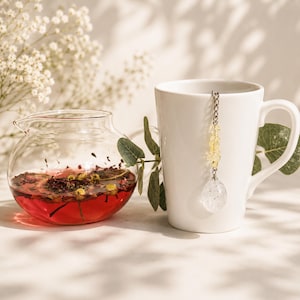 May include: A clear glass teapot filled with red herbal tea containing dried flowers and fruit, positioned next to a white ceramic mug. A metal tea infuser with a silver chain and yellow crystal beads hangs from the rim of the white mug.