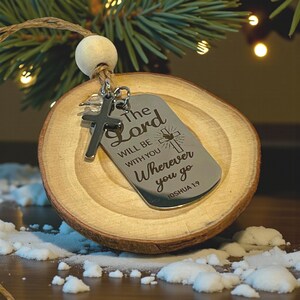 May include: A wooden Christmas ornament with a silver tag and cross charm. The tag reads "The Lord will be with you wherever you go" from Joshua 1:9. The ornament is attached to a wooden bead and twine.