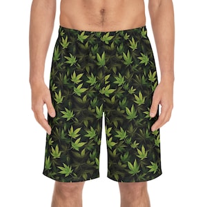 Op de afbeelding: Zwarte korte broek met een herhaald patroon van groene cannabisbladeren. De bladeren hebben verschillende tinten groen en geel, wat contrasteert met de donkere achtergrond. De korte broek is gemaakt van een lichte stof.