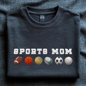 May include: Dark gray sweatshirt with the text "SPORTS MOM" in white block letters. Below the text are illustrations of a football, basketball, volleyball, rugby ball, soccer ball, and baseball.