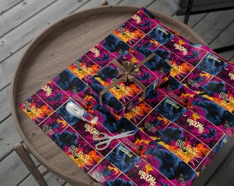 May include: A wrapped gift box and wrapping paper with a comic book design. The design features a superhero in action, with the words "Blam", "Boom", and "Kaboom" in speech bubbles. The background is pink, orange, and blue. A brown ribbon is tied around the gift box.