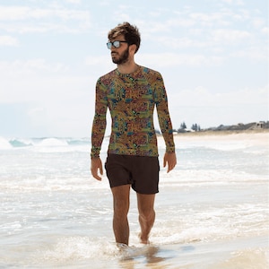 May include: A man wearing a long-sleeved shirt with a circuit board pattern and dark brown shorts. He is standing on a sandy beach with the sea in the background.