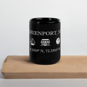 Puede incluir: Taza de café de cerámica negra con texto e iconos blancos. El texto dice "Greenport, NY" y "41.1034° N, 72.3593° W". Los iconos son un velero, una tienda y un tenedor y un cuchillo.