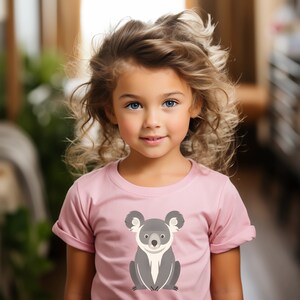 Puede incluir: Una niña con el pelo largo y rizado rubio lleva una camiseta rosa con un dibujo de un koala de dibujos animados. El koala es gris con pelaje blanco alrededor de los ojos y la nariz.