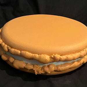 May include: A large, round, orange-brown macaron with a white filling. The macaron has a textured, ruffled edge.
