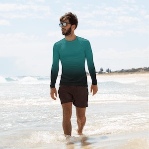 Może przedstawiać: Koszulka z długim rękawem chroniąca przed wysypką z gradientem od turkusu do czerni. Koszulka jest połączona z brązowymi szortami. Osoba spaceruje po słonecznej plaży.