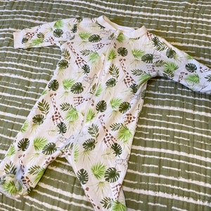 May include: White long-sleeved baby onesie with a zip closure. The onesie features a print of green leaves and brown giraffes.