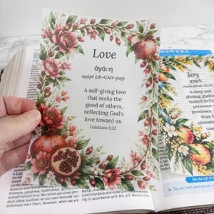 May include: A rectangular card featuring a floral border of pink flowers and pomegranates surrounding the text Love, the Greek word agape, its pronunciation, and the definition: A self-giving love that seeks the good of others.
